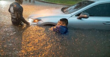 Two men help a person out of a car submerged in floodwaters.