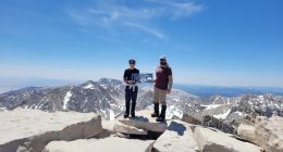 Two hikers on the summit of Mount Whitney holding a sign.