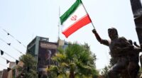 Iranian flag held aloft by a statue in Tehran, with an anti-Israel billboard in the background.