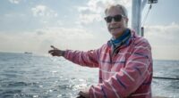 Nigel Farage on a boat, pointing towards a distant ship.