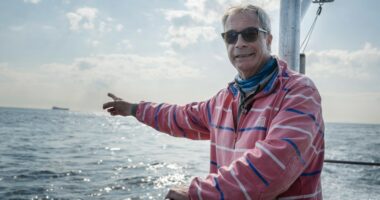 Nigel Farage on a boat, pointing towards a distant ship.