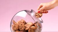 A hand taking a chocolate chip cookie from a cookie jar.