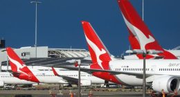 The vertical stabilisers of Qantas planes at Sydney Kingsford-Smith Airport, from the left: a Boeing B737-838 plane, registration VH-XZD; a Boeing B787-9 plane, registration VH-ZNE; and an Airbus A380-842 plane, registration VH-OQB. In the background on the left is a fourth Qantas plane, a Boeing B737-838, registration VH-XZH. In the far background is the international terminal.  In the foreground are navigational light structures at the northern end of the runway.  This image was taken from Nig