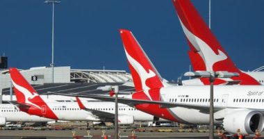 The vertical stabilisers of Qantas planes at Sydney Kingsford-Smith Airport, from the left: a Boeing B737-838 plane, registration VH-XZD; a Boeing B787-9 plane, registration VH-ZNE; and an Airbus A380-842 plane, registration VH-OQB. In the background on the left is a fourth Qantas plane, a Boeing B737-838, registration VH-XZH. In the far background is the international terminal.  In the foreground are navigational light structures at the northern end of the runway.  This image was taken from Nig