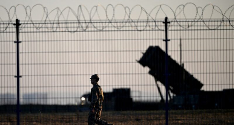 Soldier walking past Patriot air defense systems at a military hub.