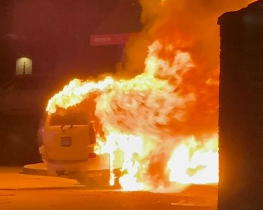 Car engulfed in flames near a building.