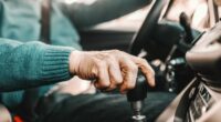 Close up of senior man holding one hand on gearshift and other on steering wheel while sitting in his car. Selective focus on hand.