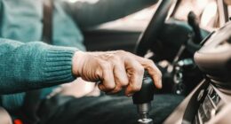 Close up of senior man holding one hand on gearshift and other on steering wheel while sitting in his car. Selective focus on hand.