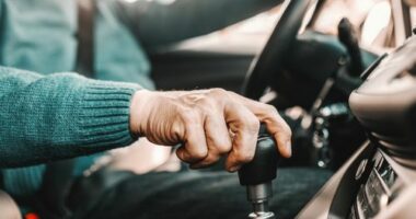 Close up of senior man holding one hand on gearshift and other on steering wheel while sitting in his car. Selective focus on hand.