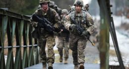 Two soldiers carrying rifles walk across a metal bridge.