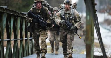 Two soldiers carrying rifles walk across a metal bridge.