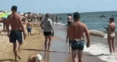 Crowded beach with people walking on the sand and in the water.