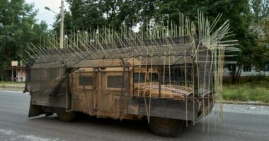 Military vehicle with cage armor driving down a road.