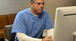 Erik Menendez with gray hear before a computer, wearing prison clothes.