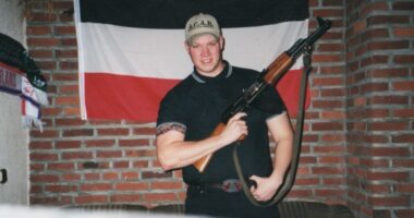 Photo of a young man holding a rifle, standing in front of a German flag.
