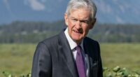 Jay Powell walks outdoors in a suit and purple tie, with greenery and mountains in the background.