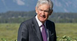 Jay Powell walks outdoors in a suit and purple tie, with greenery and mountains in the background.