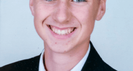 Photo of a smiling boy in a suit and red tie.