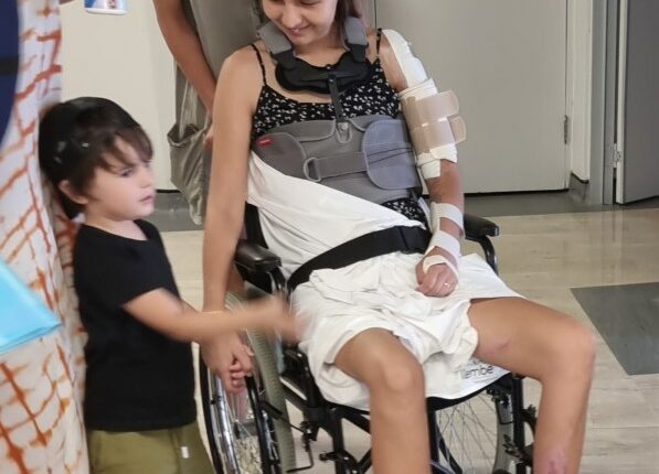 Woman in wheelchair with neck brace and arm sling, surrounded by family.
