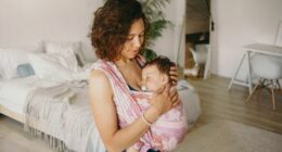Casually dressed young mixed race woman with wavy hair spending day at home, cradling her infant child, wearing baby carrier. Hispanic mother watching her adorable son sleeping in wrap sling