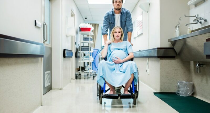 Pregnant woman in a wheelchair being pushed by a man down a hospital corridor.