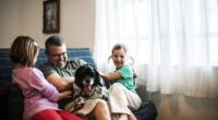 Young girls play with their father and pet the dog sitting on his lap.