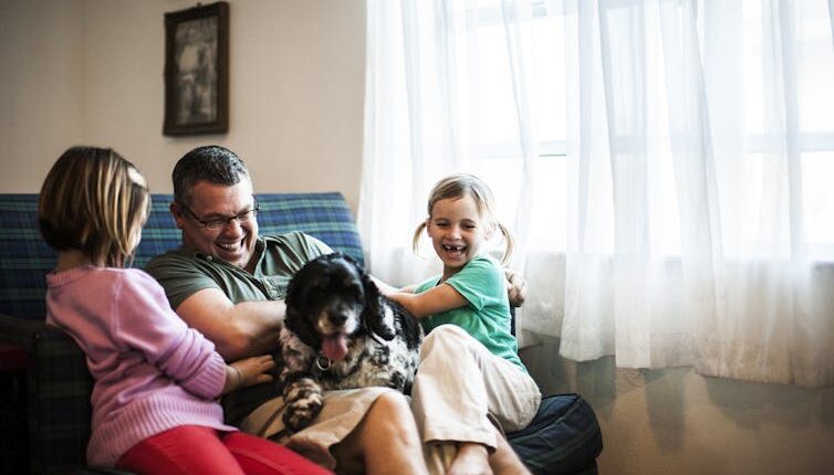 Young girls play with their father and pet the dog sitting on his lap.