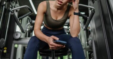 A dedicated young woman seated in the gym, deeply focused on her phone, showcasing the blend of concentration and relaxation during workout routines.