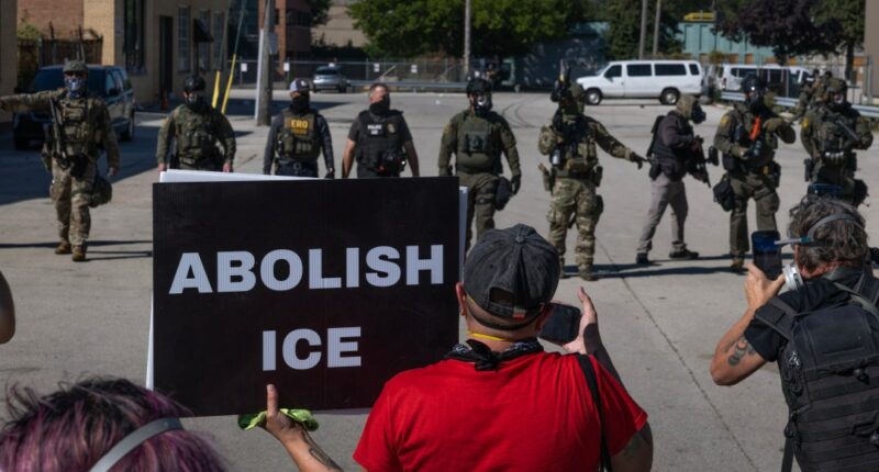 Protesters gather in front of ICE officers. One holds a sign reading ABOLISH ICE