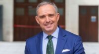 Mark Sedwill smiles while standing outdoors in a blue suit and green tie.