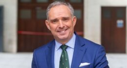 Mark Sedwill smiles while standing outdoors in a blue suit and green tie.