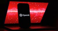 The OpenAI logo is displayed on a mobile phone screen in front of a laptop showing red digital graphics with numbers.