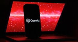 The OpenAI logo is displayed on a mobile phone screen in front of a laptop showing red digital graphics with numbers.