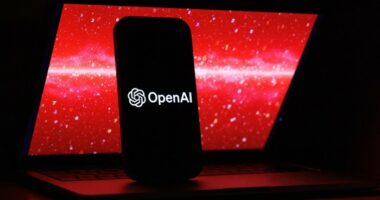 The OpenAI logo is displayed on a mobile phone screen in front of a laptop showing red digital graphics with numbers.