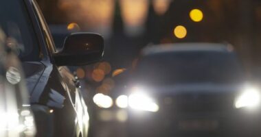 Cars in traffic during dusk, showcasing headlights and city lights, with a blurred background reflecting urban life