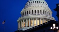 The dome of the US Capitol is illuminated at dusk with an American flag and street lamps visible in the foreground.