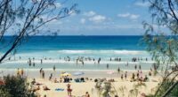 Horizontal Seascape of people enjoying the summer sun on busy tourist beach holiday with sand turquoise waves blue sky at famous surf ocean Byron Bay Australia