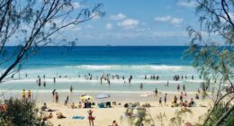 Horizontal Seascape of people enjoying the summer sun on busy tourist beach holiday with sand turquoise waves blue sky at famous surf ocean Byron Bay Australia