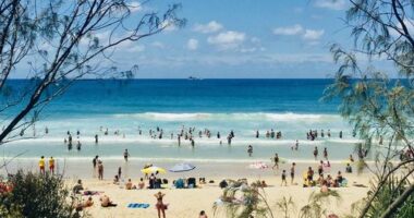 Horizontal Seascape of people enjoying the summer sun on busy tourist beach holiday with sand turquoise waves blue sky at famous surf ocean Byron Bay Australia