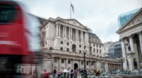 The Bank of England building with a blurred red double-decker bus passing in front and people gathered outside.