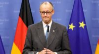 Friedrich Merz stands at a podium during a media conference, with German and EU flags in the background.