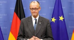 Friedrich Merz stands at a podium during a media conference, with German and EU flags in the background.