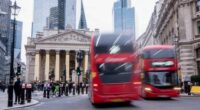 Red double-decker buses pass in front of the Bank of England, with people walking and a cyclist nearby in central London.