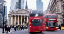 Red double-decker buses pass in front of the Bank of England, with people walking and a cyclist nearby in central London.