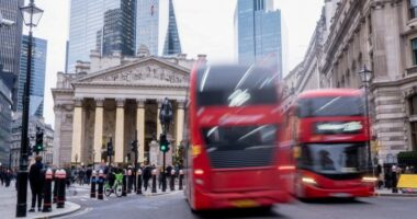 Red double-decker buses pass in front of the Bank of England, with people walking and a cyclist nearby in central London.