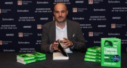 Stephen Witt sits at a table with stacks of his book, ‘The Thinking Machine’, preparing to sign a copy.