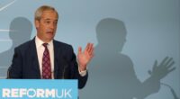 Nigel Farage speaks at a podium labeled &quot;Reform UK&quot;, gesturing with his hand during a conference.