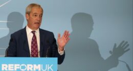 Nigel Farage speaks at a podium labeled &quot;Reform UK&quot;, gesturing with his hand during a conference.