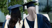 Two female graduates in academic gowns and caps stand together outdoors, one holding flowers, at Birmingham City University graduation day.