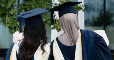 Two female graduates in academic gowns and caps stand together outdoors, one holding flowers, at Birmingham City University graduation day.